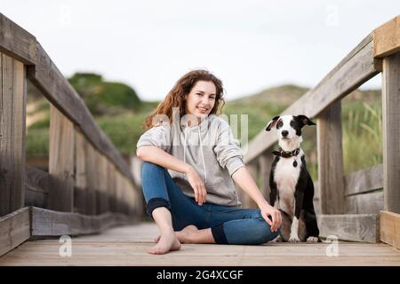 Lächelnde junge Frau, die von Jack Russell Terrier Hund auf einer Holzbrücke sitzt Stockfoto