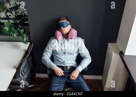 Geschäftsmann mit Augenmaske und Nackenkissen, die auf einem Bürostuhl ruhen Stockfoto