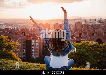 Frau mit erhobenen Armen und Blick auf die Stadt, während sie während des Sonnenuntergangs im Park sitzt Stockfoto