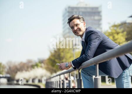 Lächelnder männlicher Unternehmer, der sich an sonnigen Tagen auf Geländer stützt Stockfoto