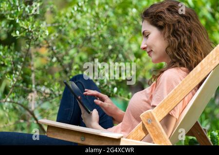 Junge Frau, die im Garten auf einem Stuhl sitzt, mit einem digitalen Tablet Stockfoto