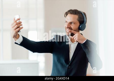 Geschäftsmann mit Kopfhörern, die zeigen, während er im Büro Selfie über das Smartphone nimmt Stockfoto