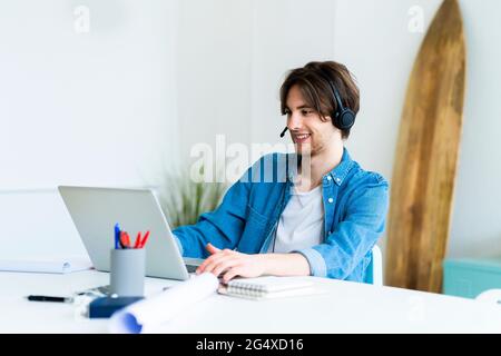Männlicher Unternehmer mit Headset, der während der Arbeit im Büro einen Laptop verwendet Stockfoto