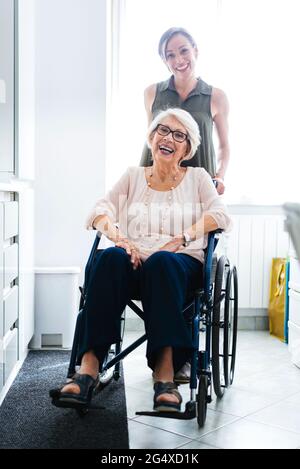 Fröhliche behinderte ältere Frau, die auf dem Rollstuhl sitzt, von der Enkelin, die zu Hause in der Küche steht Stockfoto