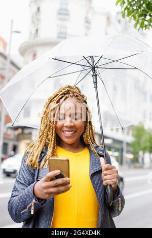 Lächelnde Frau, die Smartphone benutzt, während sie den Regenschirm hält Stockfoto