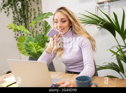 Lächelnde blonde Geschäftsfrau, die über einen Lautsprecher im Büro auf dem Mobiltelefon spricht Stockfoto