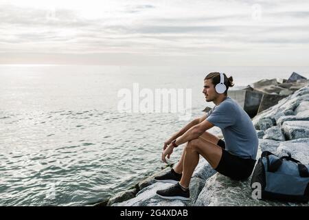 Sportler mit Kopfhörern, die die Sicht betrachten, während sie auf Felsen am Meer sitzen Stockfoto