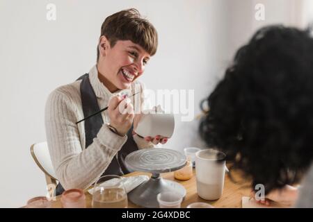 Lächelnde Malerin mit Pinsel und Becher, die Kollegen in der Werkstatt ansieht Stockfoto