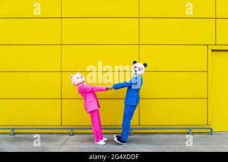 Mann und Frau tragen lebhafte Anzüge und tragen Masken, die die Hände an der gelben Wand halten Stockfoto