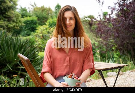 Frau mit braunem Haar hält eine Obstschale, während sie auf einem Stuhl im Garten sitzt Stockfoto