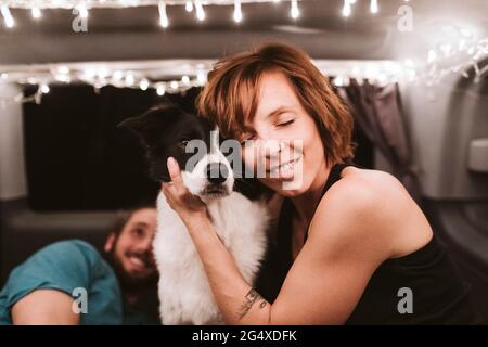 Lächelnde Frau umarmt Hund, während sie in beleuchtetem Van in der Nacht reist Stockfoto