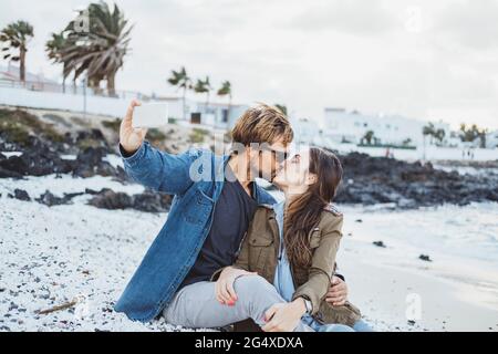 Freund küsst Freundin, während Selfie durch Smartphone am Strand Stockfoto