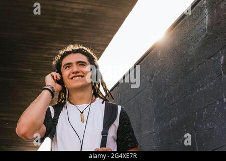 Lächelnder junger Mann, der wegschaut, während er Musik über Kopfhörer an der Wand hört Stockfoto