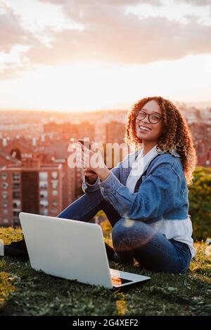 Lächelnde Frau mit Smartphone und Laptop sitzt im Park während des Sonnenuntergangs Stockfoto