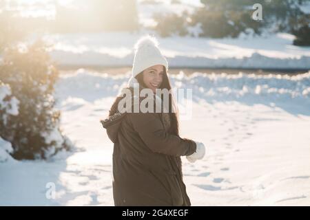 Lächelnde Frau, die während eines sonnigen Tages auf Schnee steht und über die Schulter blickt Stockfoto