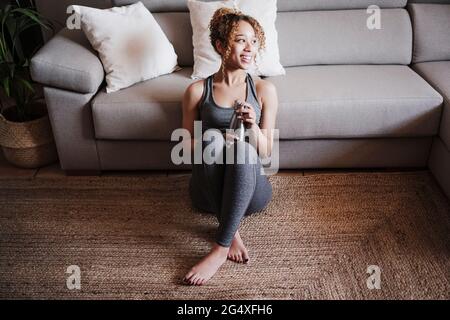 Lächelnde junge Frau, die zu Hause auf dem Teppich vor dem Sofa sitzt Stockfoto