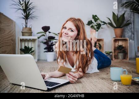Lächelnde Rothaarige Frau mit Kreditkarte beim Online-Shopping auf dem Laptop zu Hause Stockfoto