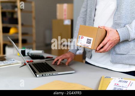 Ein junger Geschäftsmann hält einen Pappkarton in der Hand, während er einen Laptop am Schreibtisch benutzt Stockfoto