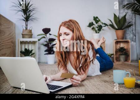 Frau mit Kreditkarte macht Online-Shopping über Laptop, während sie zu Hause auf dem Boden liegt Stockfoto