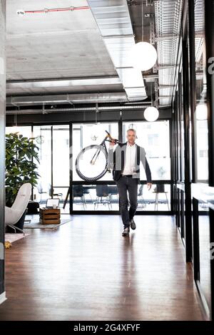 Geschäftsmann, der Fahrrad auf der Schulter trägt, während er im Büro läuft Stockfoto