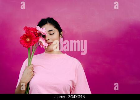 Junge Frau, die vor der Wand die Augen mit Gerbera-Gänseblümchen bedeckt Stockfoto