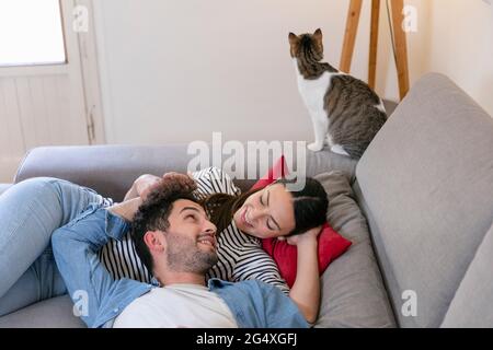 Lächelndes Paar mit tabby Katze, die auf dem Sofa im Wohnzimmer im Zimmer liegt Stockfoto