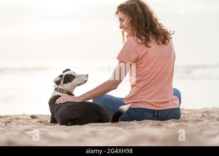 Lächelnde junge Frau streichelte Jack Russell Terrier, während sie am Strand saß Stockfoto