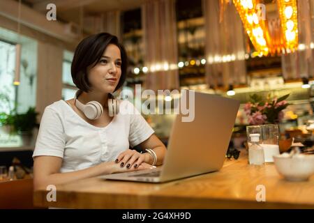 Lächelnde junge Freiberuflerin, die mit einem Laptop im Restaurant sitzt und wegschaut Stockfoto