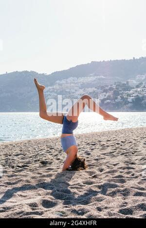 Frau mit mittlerem Erwachsenenalter, die bei Sonnenschein am Strand Kopfstand übt Stockfoto