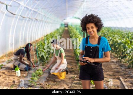 Lächelndes afro-Mädchen mit digitalem Tablet, das am Gewächshaus steht, während Farmerinnen im Hintergrund arbeiten Stockfoto