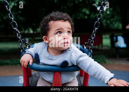 Kleiner Junge schaut weg, während er auf der Schaukel auf dem Spielplatz sitzt Stockfoto