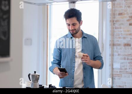 Junger Mann mit Mobiltelefon, während er in der heimischen Küche eine Kaffeetasse hält Stockfoto