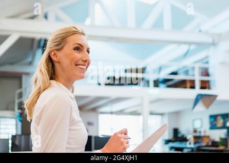 Lächelnde blonde Frau mit Dokumenten im kreativen Büro, die wegschauen Stockfoto