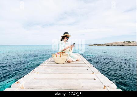 Lächelnde Frau, die während des Urlaubs auf dem Steg saß, mit dem Mobiltelefon Stockfoto