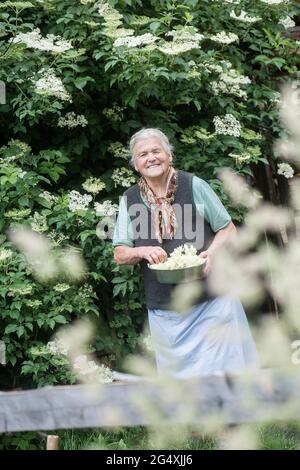 Lächelnde ältere Frau mit Holunderblüten vor dem Hof Stockfoto