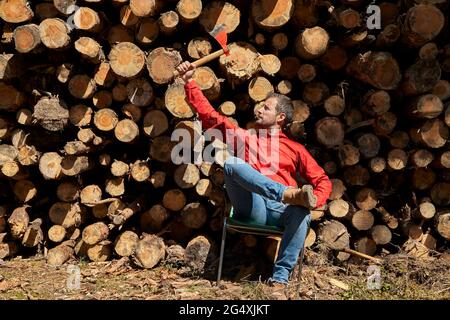 Männlicher Logger, der Axt hält, während er in der Holzindustrie vor Baumstämmen sitzt Stockfoto