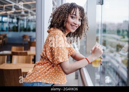 Lächelnde junge Frau mit Einweg-Teetasse, die auf dem Geländer am Balkon im Café gelehnt ist Stockfoto