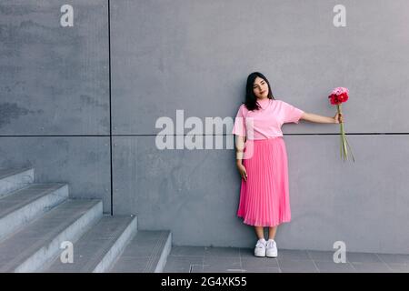 Junge Frau schaut weg, während sie rosa Blumen vor der grauen Wand hält Stockfoto