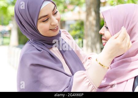 Lächelnde junge Frau, die den Hijab einer Freundin im Park anpasst Stockfoto