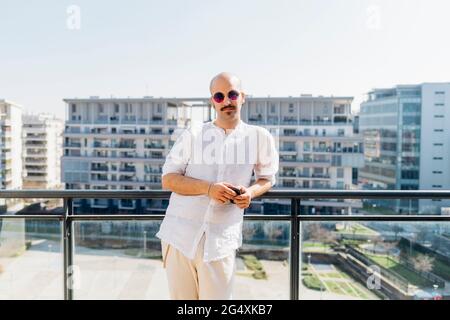 Junger Mann mit Sonnenbrille, der sich an sonnigen Tagen auf dem Balkon am Geländer stützt Stockfoto