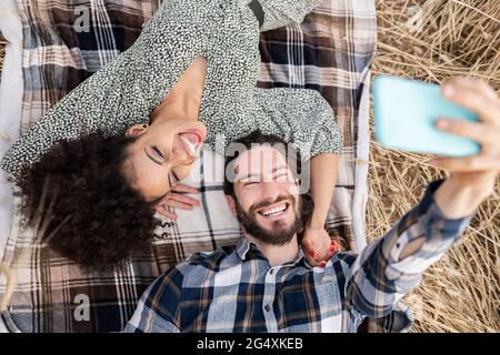 Fröhlicher Freund, der Selfie über das Smartphone mit Frau macht, während er auf der Decke liegt Stockfoto
