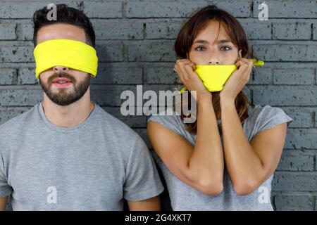 Freundin bedeckt den Mund mit einem Tuch, während sie vor der Wand neben einem Freund mit verbundenen Augen steht Stockfoto