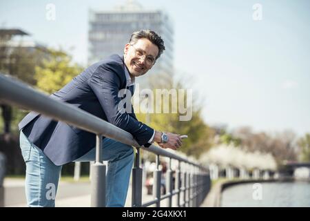 Reifer Geschäftsmann lächelt, während er sich auf das Geländer stützt Stockfoto