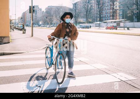 Junge Frau mit Gesichtsmaske und Hut zu Fuß mit dem Fahrrad auf der Straße in der Stadt Stockfoto