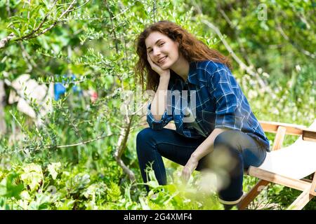 Lächelnde junge Frau mit der Hand am Kinn, die im Garten auf einem Stuhl sitzt Stockfoto