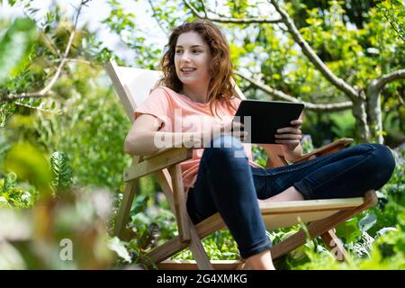 Junge Frau mit digitalem Tablet schaut weg, während sie auf einem Stuhl im Garten sitzt Stockfoto