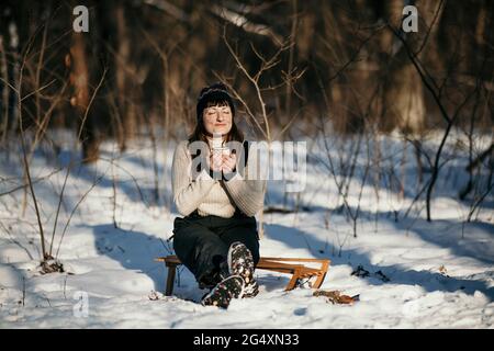 Frau hält den Becher, während sie im Wald auf dem Schlitten sitzt Stockfoto