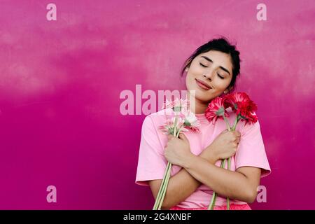 Junge Frau mit gekreuzten Armen, die einen Bund Gerbera-Gänseblümchen vor einer rosa Wand hält Stockfoto