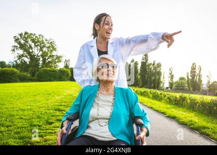 Fröhliche weibliche Hausmeisterin zeigt, während sie eine ältere Frau im Park mit Rollen Stockfoto