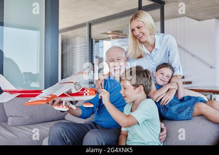 Älterer Mann, der ein Spielzeug-Flugzeug hält und mit einem Jungen spielt, während er von Tochter und Mädchen im Wohnzimmer sitzt Stockfoto